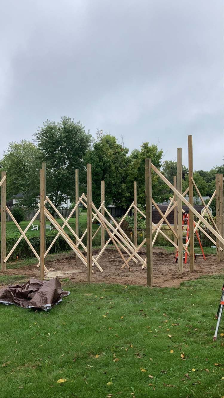 Wooden structure under construction in a yard. Tall posts with X-bracing. Cloudy sky.