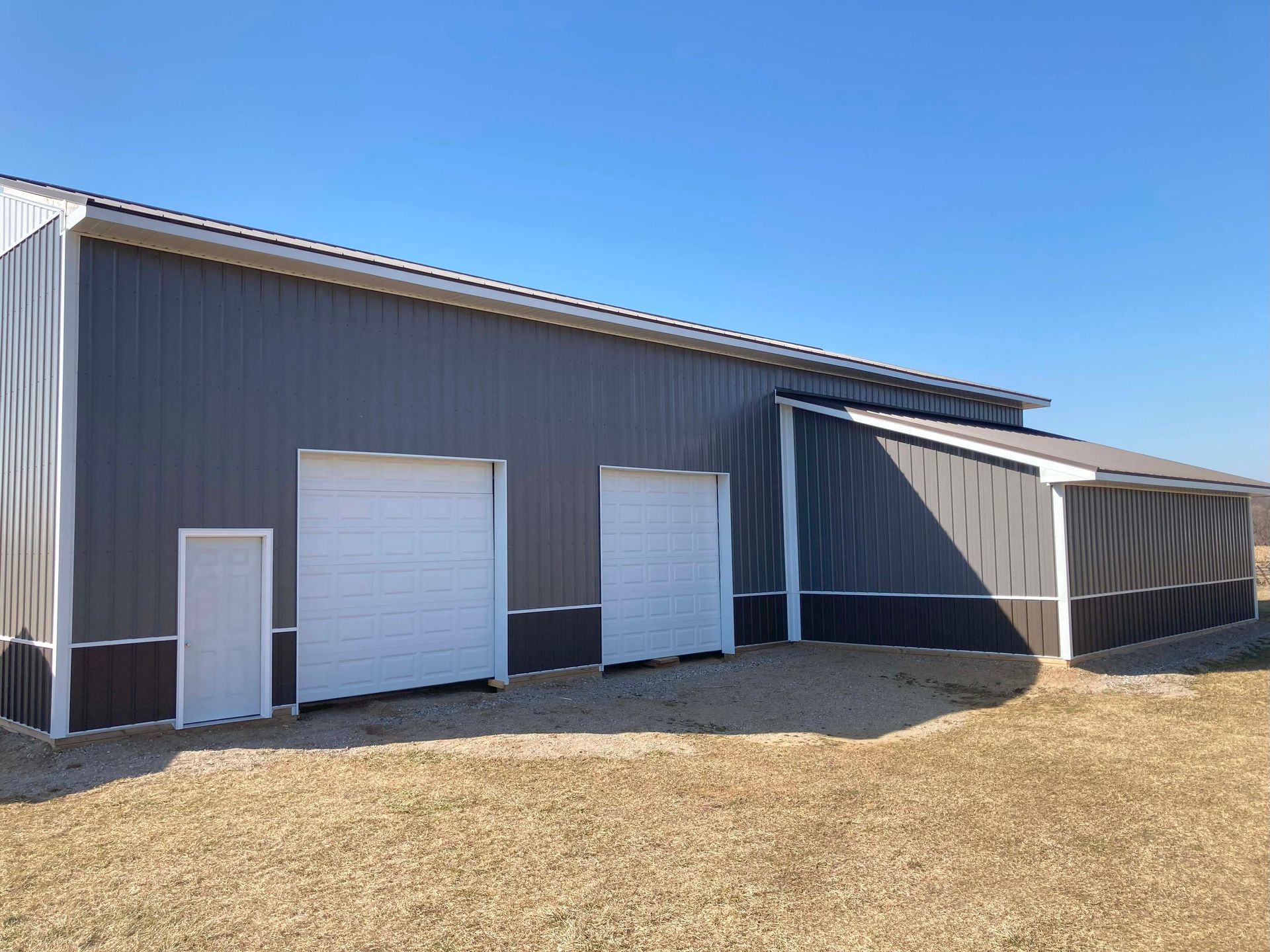 Gray and brown metal barn with white doors under a clear blue sky.