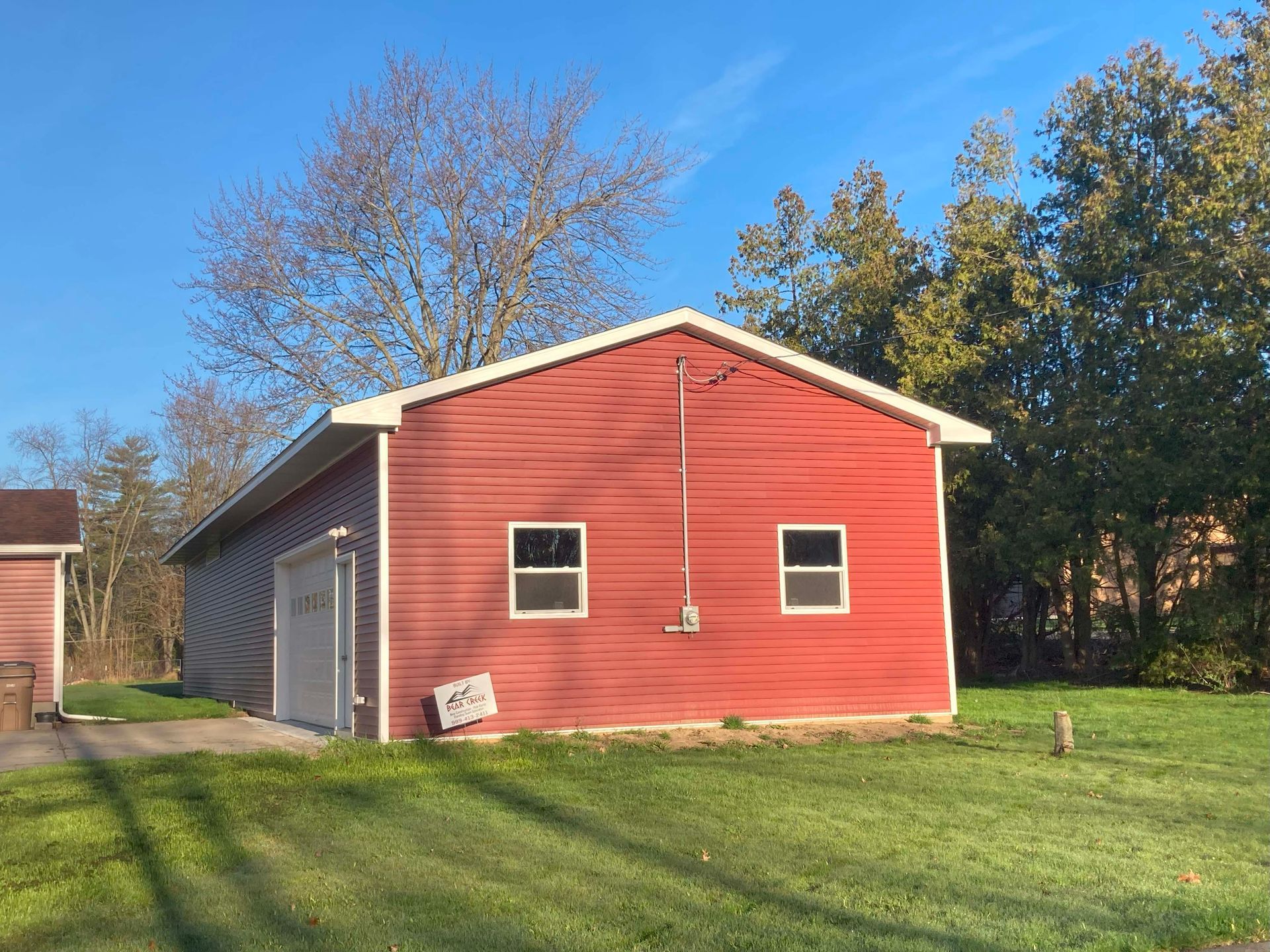 Red building with a garage door and two windows on a grassy lawn with trees and blue sky.