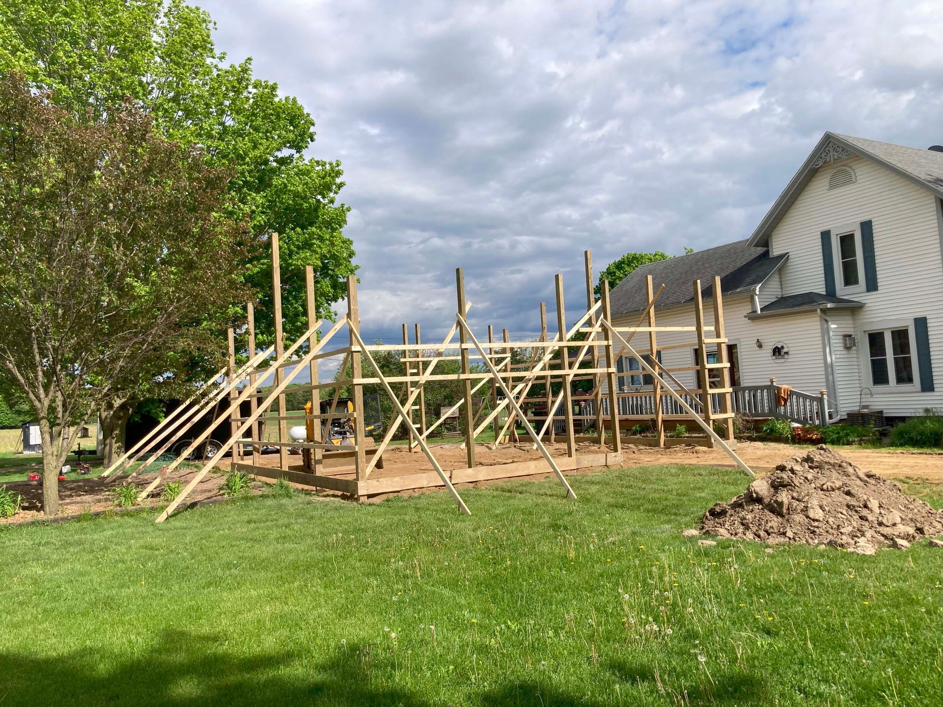 Wooden frame construction for a new building in a grassy yard, near a house on a cloudy day.