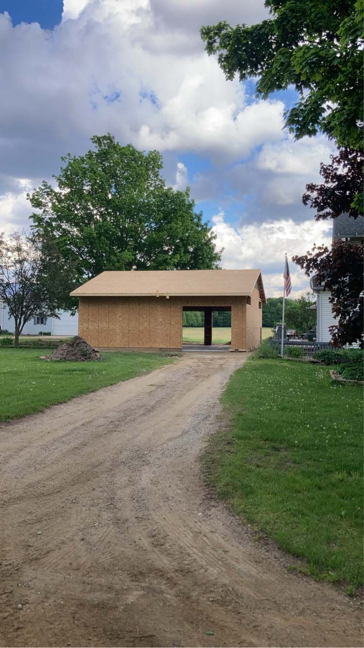 Gravel driveway leading to a partially-built tan wooden building with a green lawn and trees under a cloudy sky.