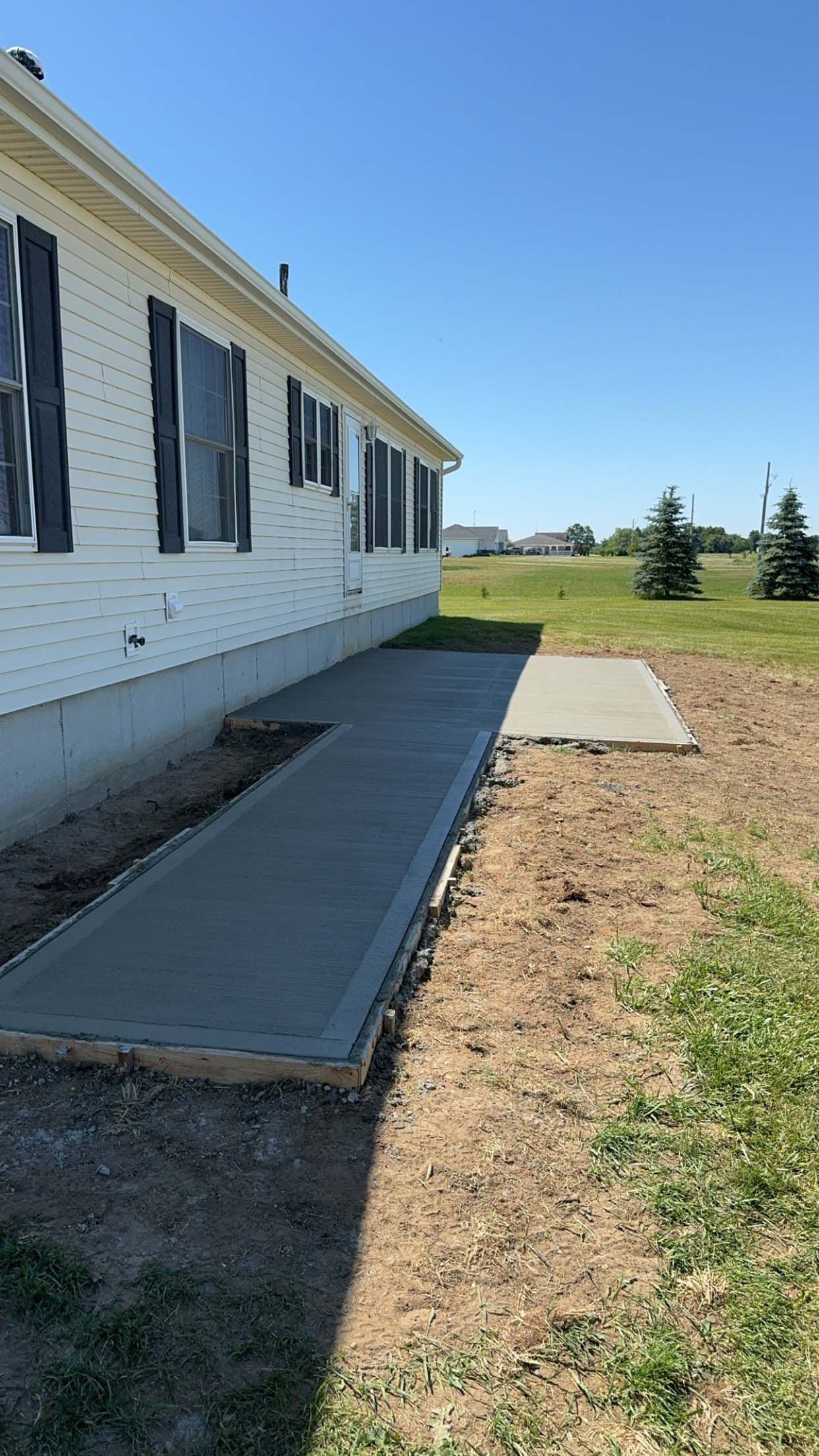 Newly poured concrete walkway next to a white house with black shutters on a sunny day.
