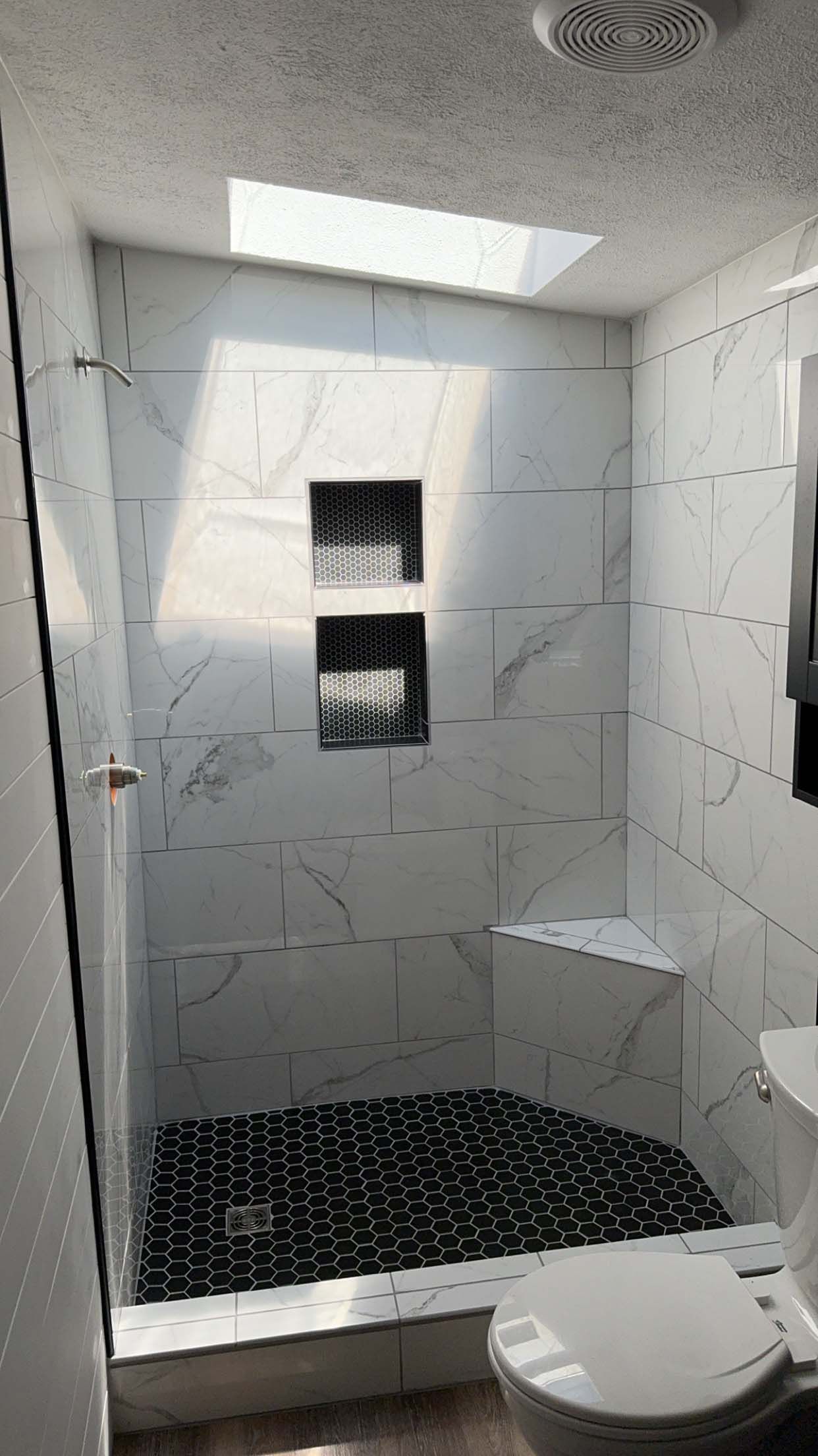 Modern shower with marble-like tiles, a pebbled floor, built-in shelving, and a skylight.