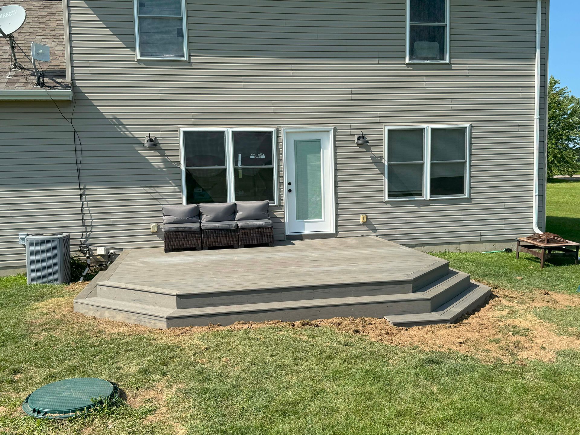 Back of house with a gray deck, steps, and a couch. Grass and a clear blue sky.