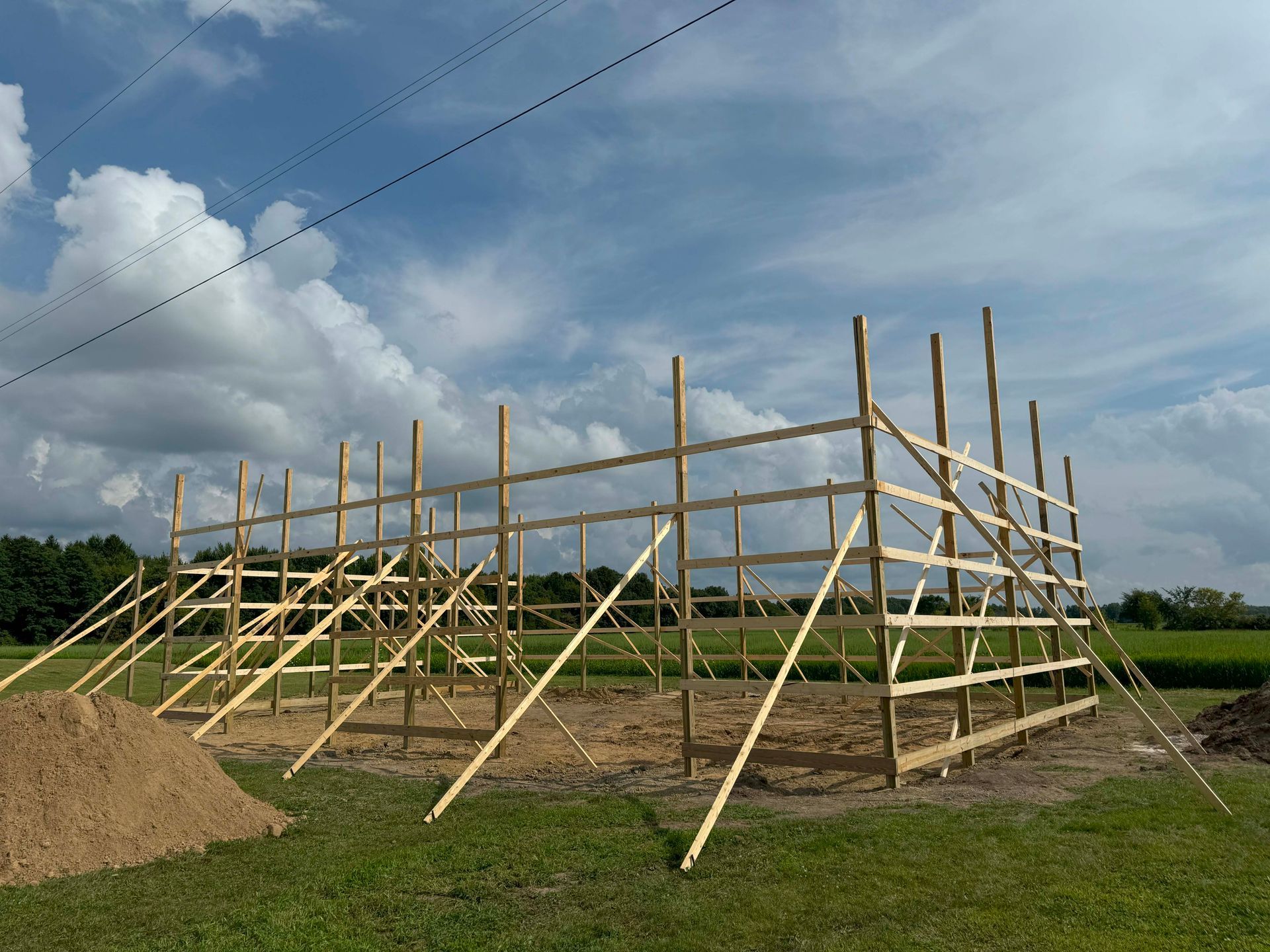 A wooden barn frame under construction in a grassy field, with a cloudy sky above.