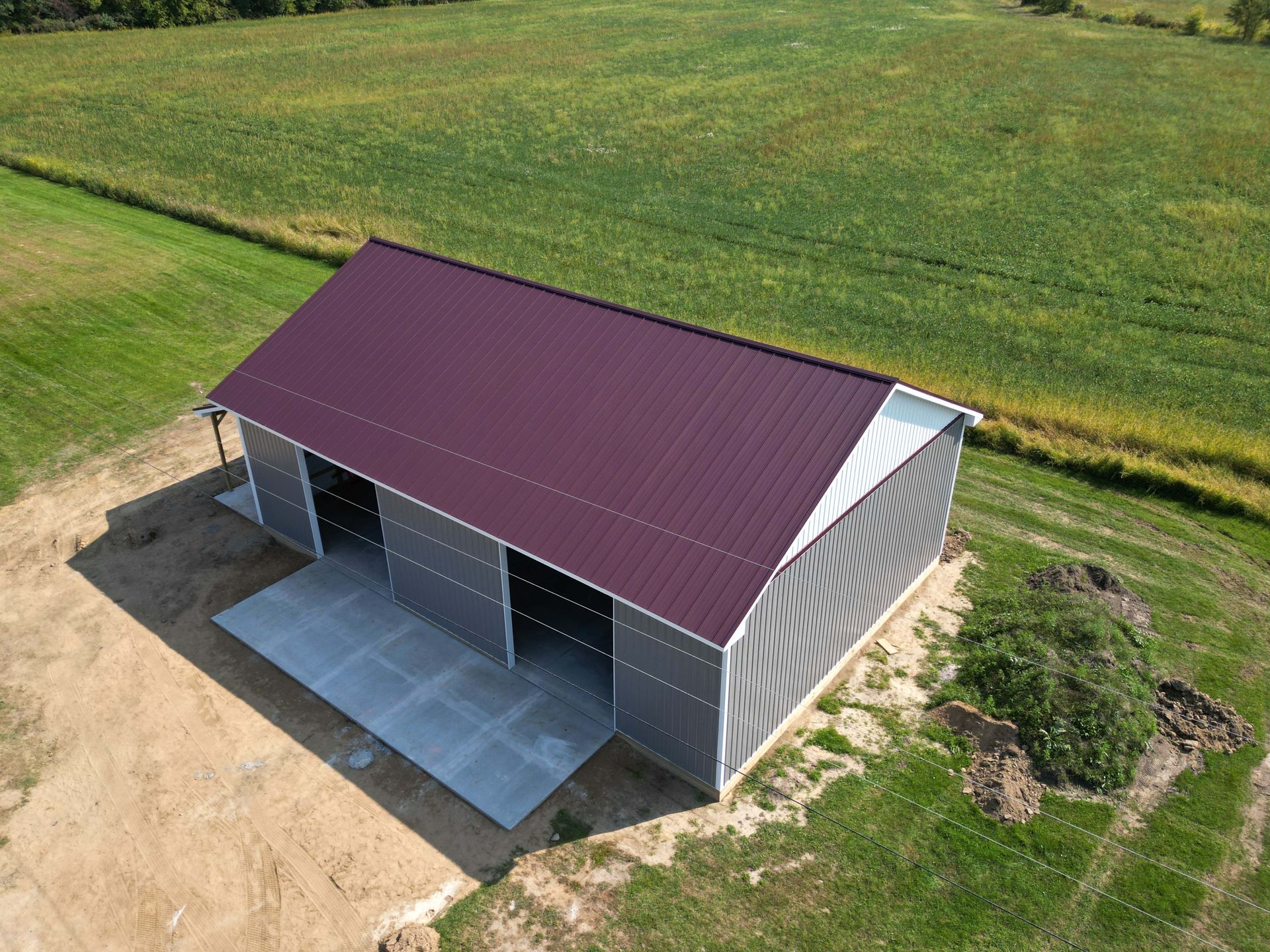 Metal barn with maroon roof, open doors, and concrete pad on a grassy field.