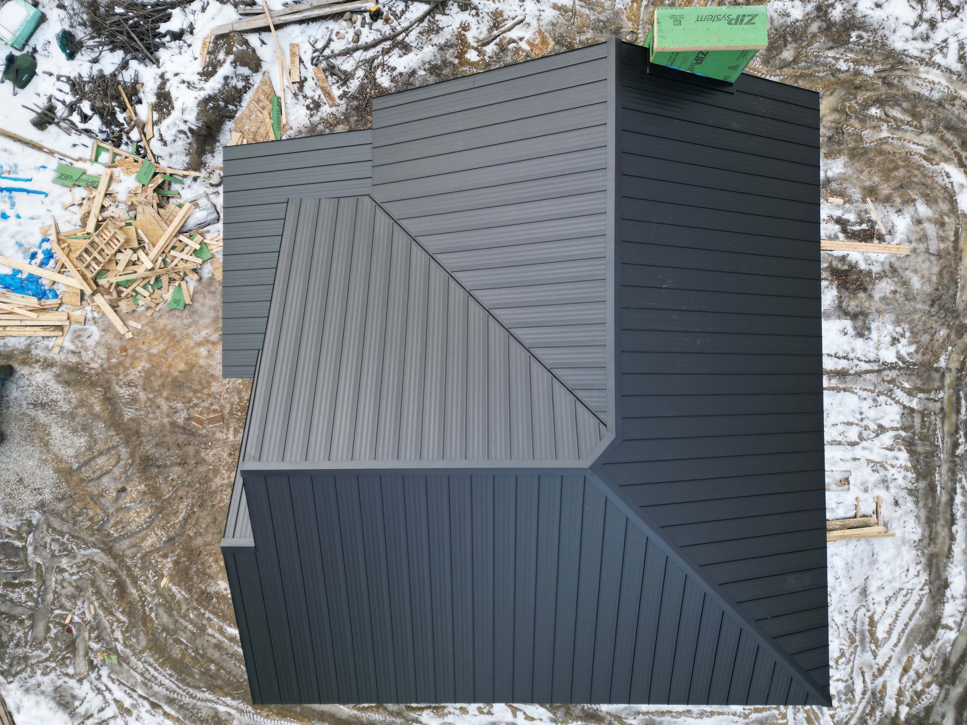 Overhead view of a house with a dark gray roof surrounded by snow and construction materials.