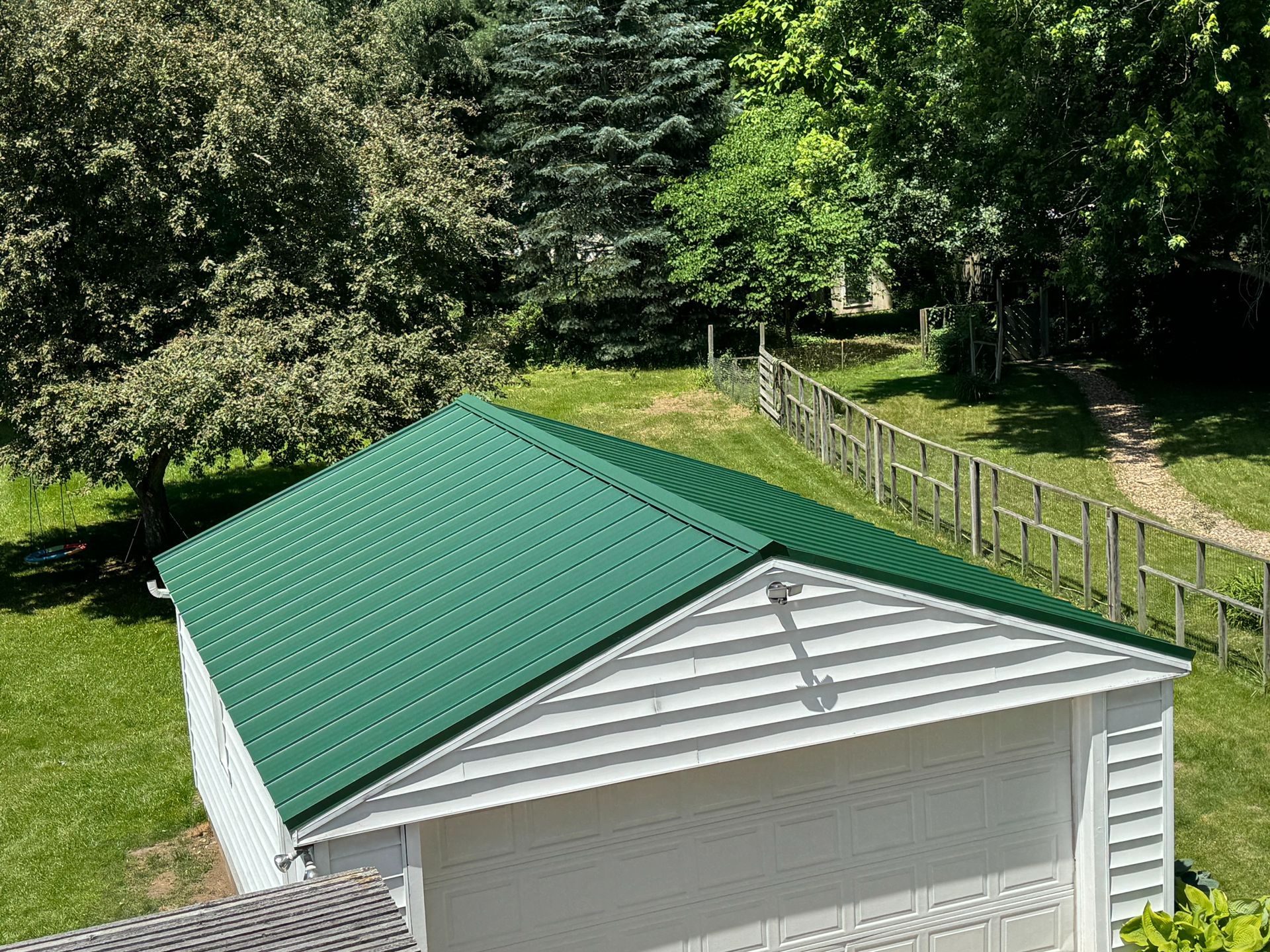 Green metal roof on a white building, surrounded by green trees and grass on a sunny day.