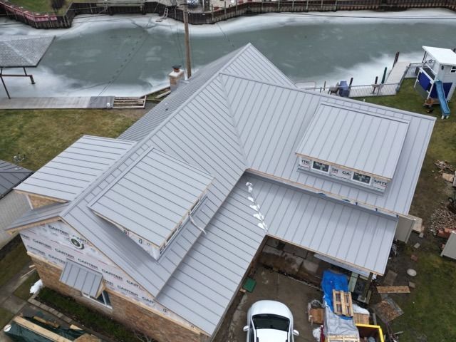 Gray metal roof on a house by a body of water, partially covered in ice.