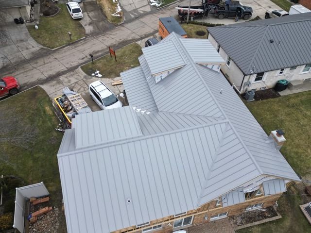 Overhead view of a house with a gray metal roof, construction materials, and vehicles in a residential area.
