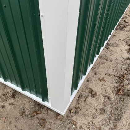 Corner of a green corrugated metal building with white trim. It rests on sandy ground.