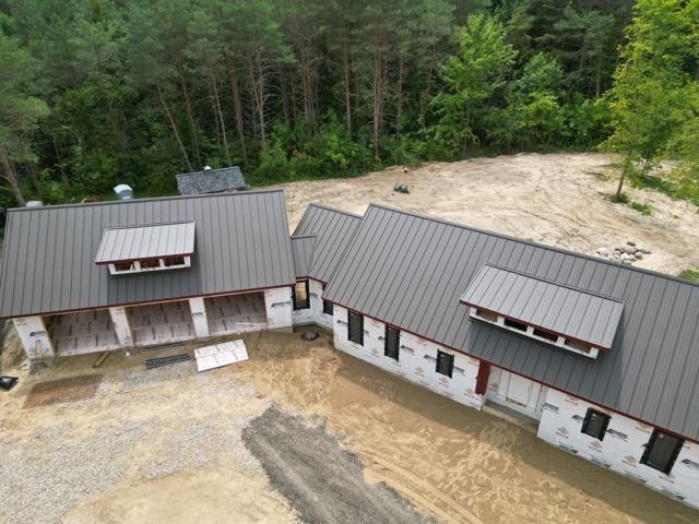 Aerial view of a partially constructed house with gray metal roof, surrounded by dirt and trees.