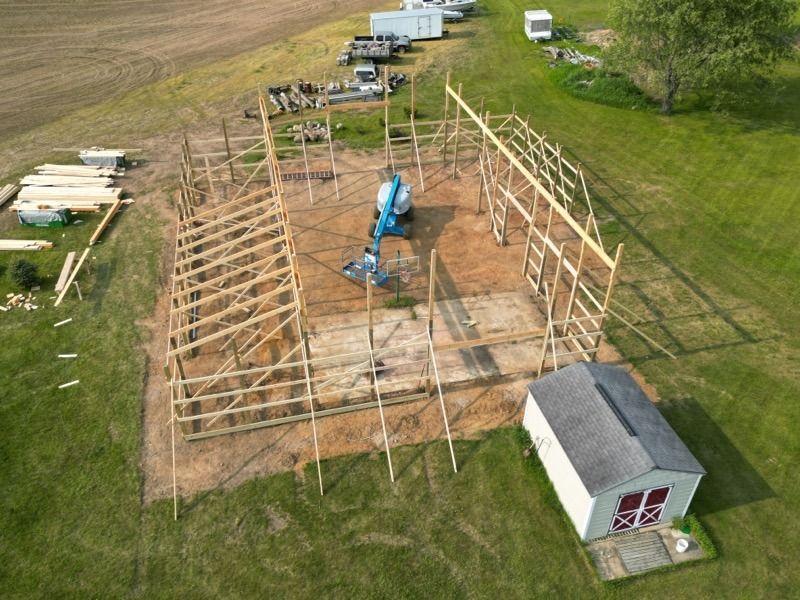 Construction site: wooden barn frame partially built on grassy field, shed in foreground, equipment nearby.