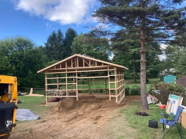 Wooden shed under construction in a yard. Blue sky, green trees, and a small pile of dirt.
