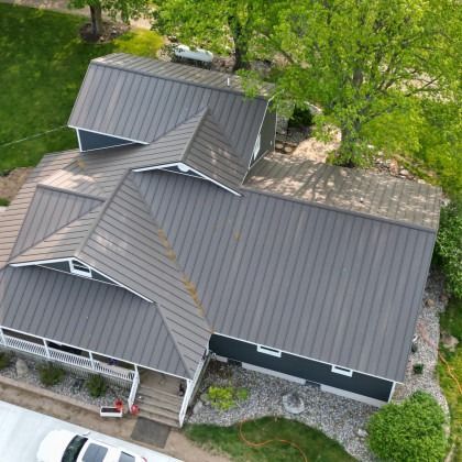 Aerial view of a house with a dark gray metal roof, surrounded by green trees and grass.