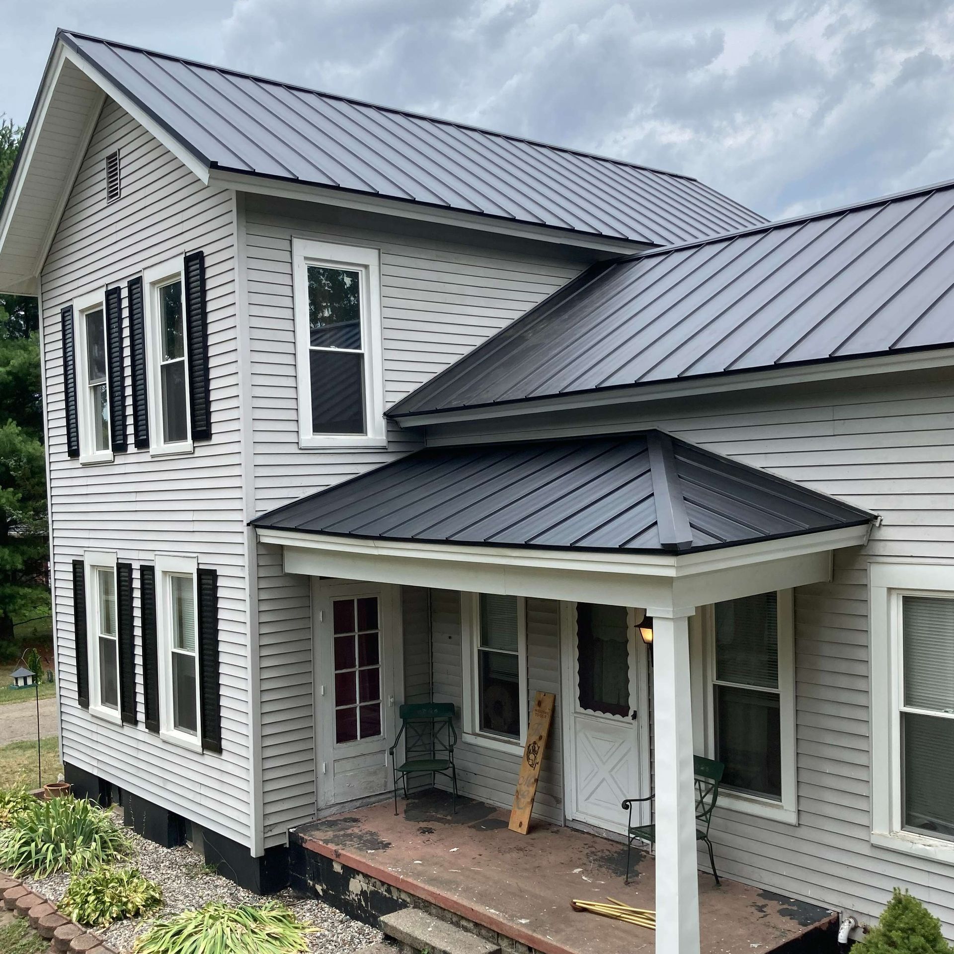 Gray house with black metal roof and shutters. Porch with chairs. Overcast sky.