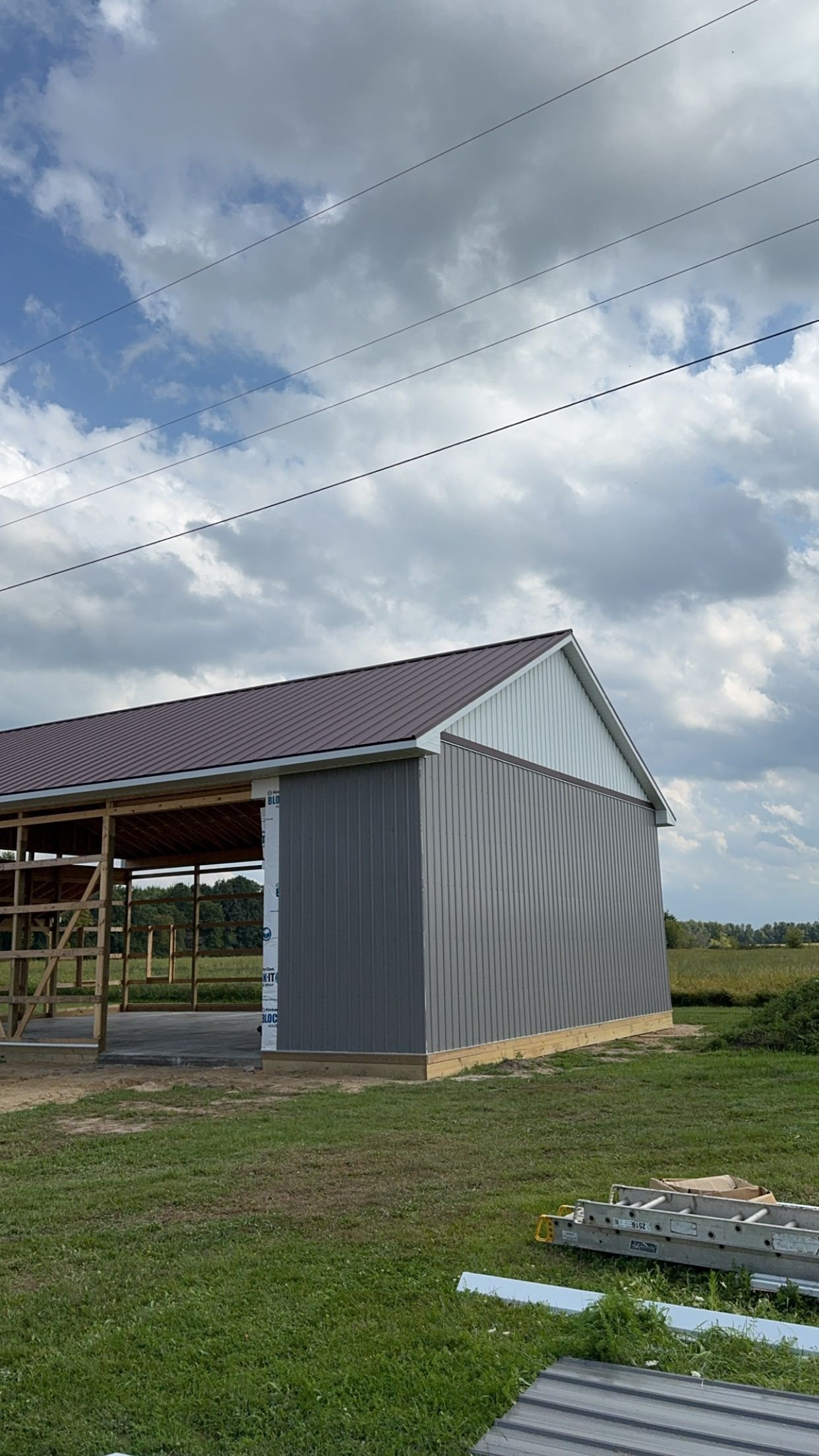 A partially constructed gray metal building with a brown roof on a grassy field under a cloudy sky.