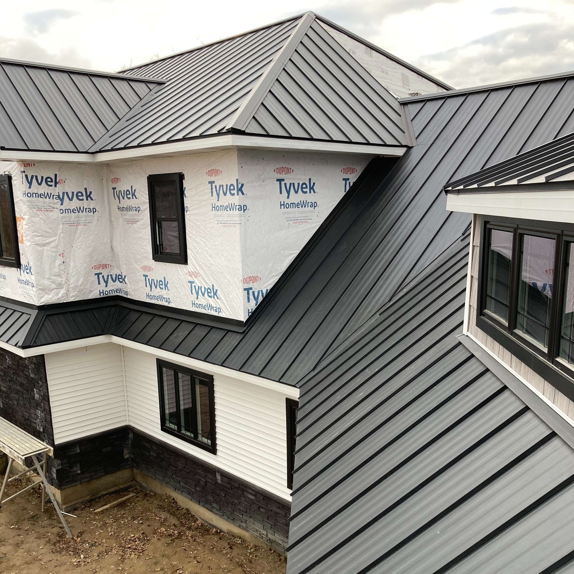 Dark gray metal roof on a house under construction; white Tyvek wrap, black window frames.