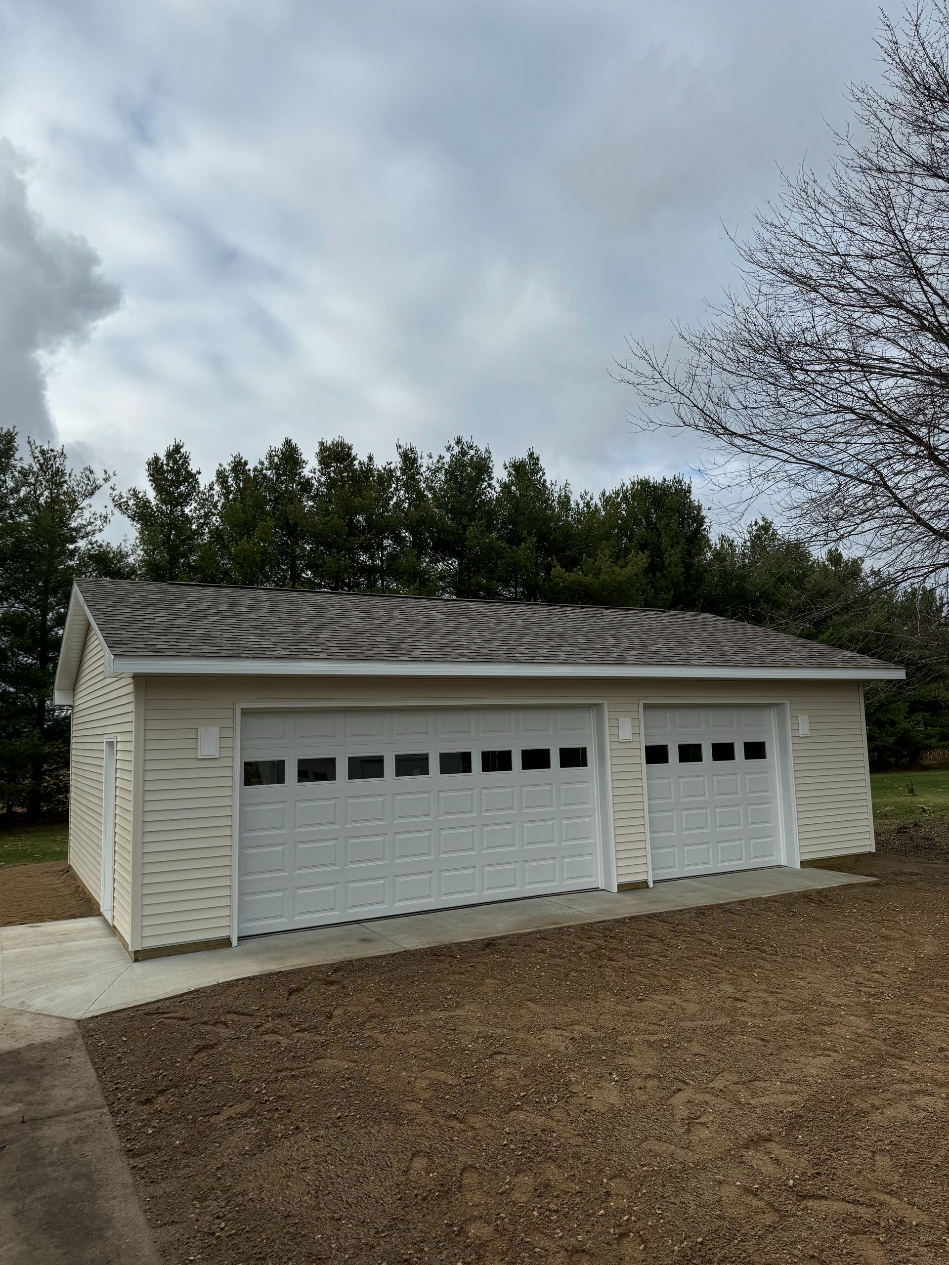 Beige garage with two white garage doors, set on concrete, with trees in the background.