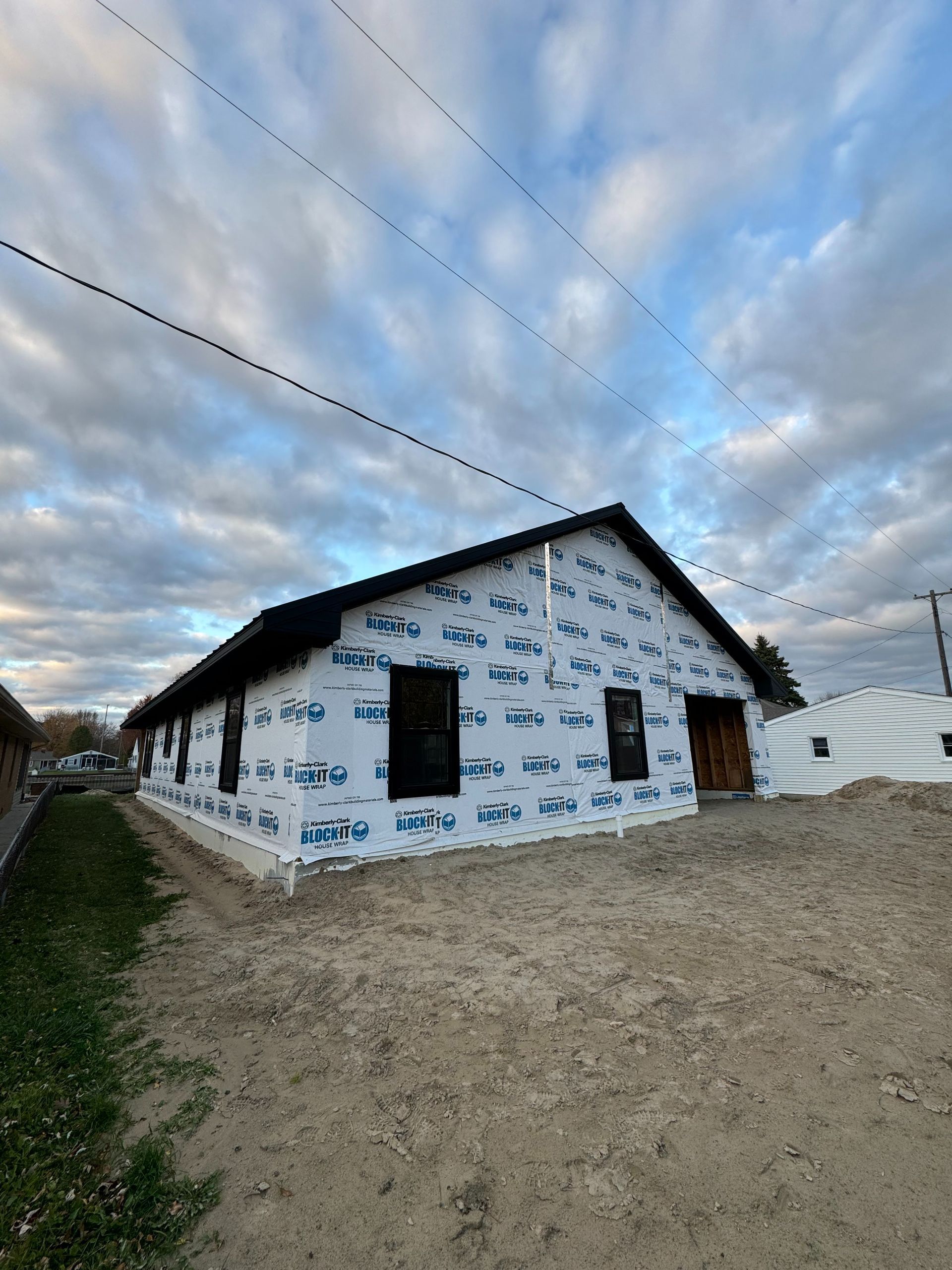 Construction of a house with black windows and roof, wrapped in blue protective sheathing, against a cloudy sky.