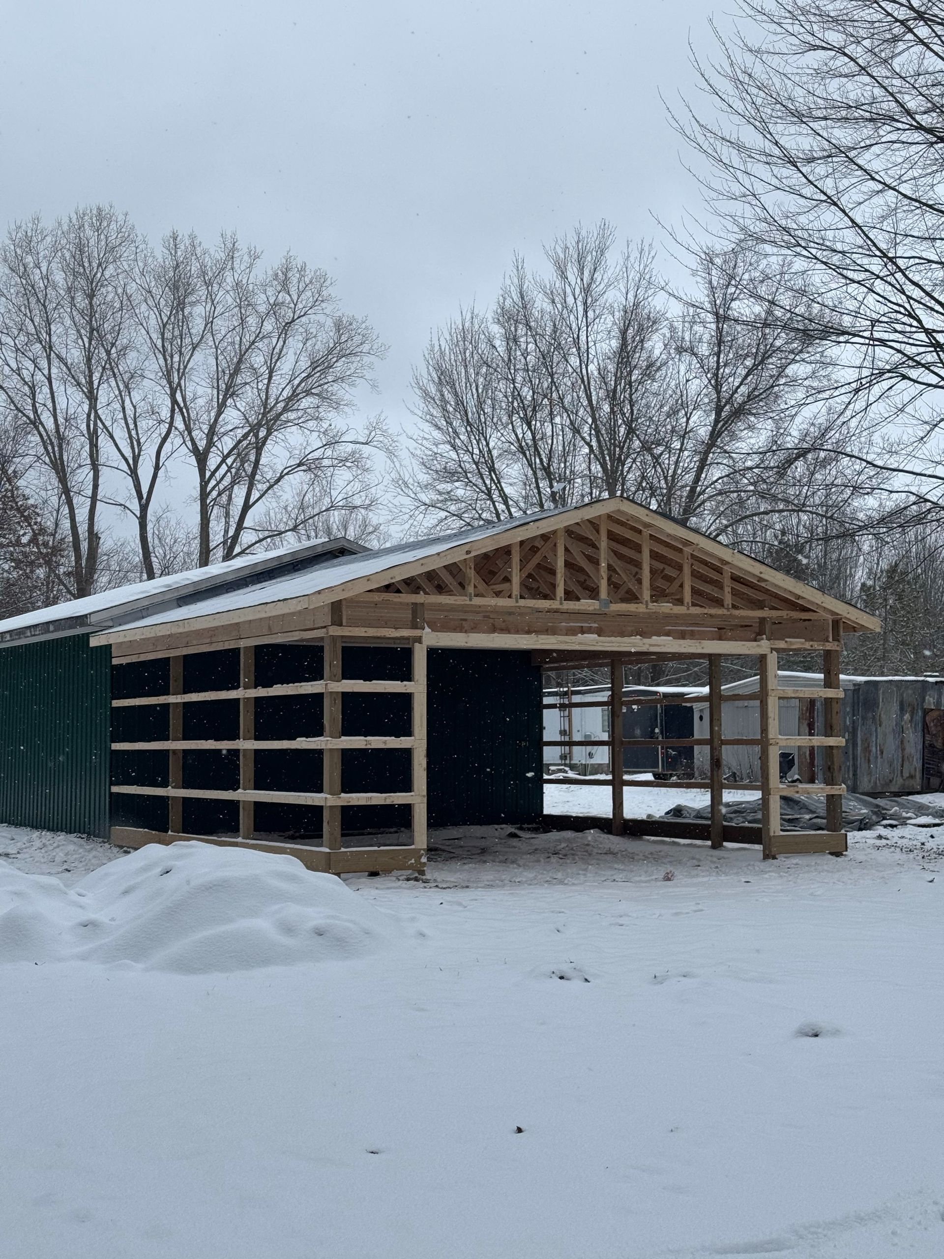 Open-sided wooden structure under construction, covered with a corrugated metal roof, in a snowy outdoor setting.