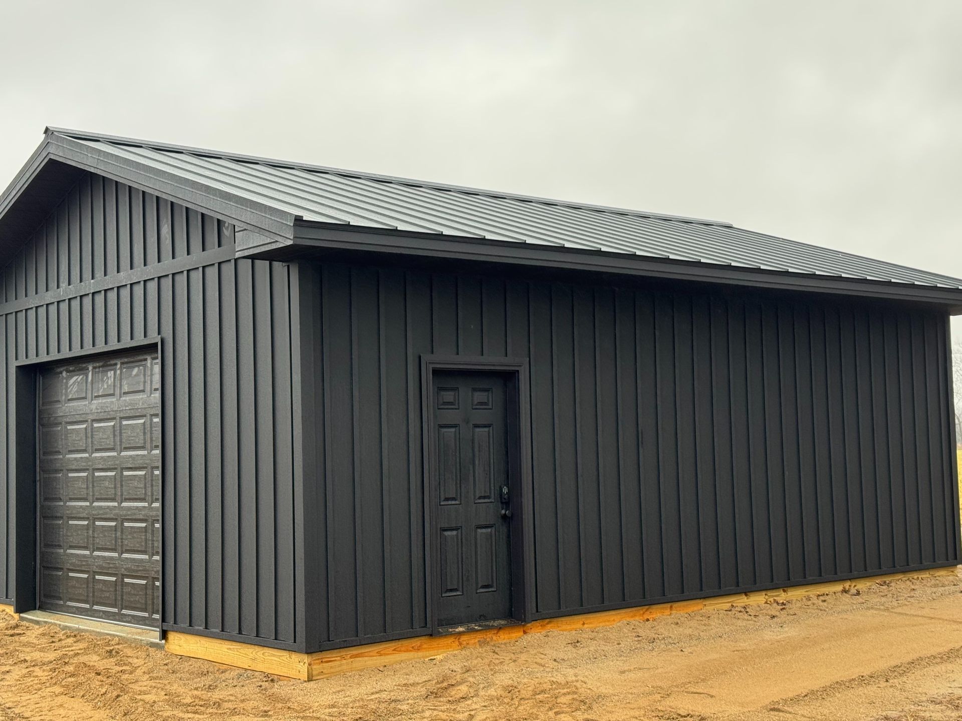 Black shed with a garage door and side door, black metal roof, in a dirt setting.