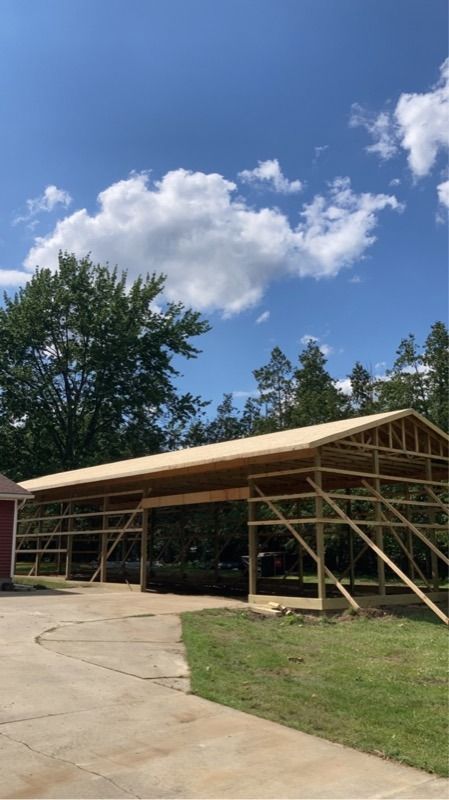A partially constructed wooden barn under a blue sky, set on concrete with green grass and trees.