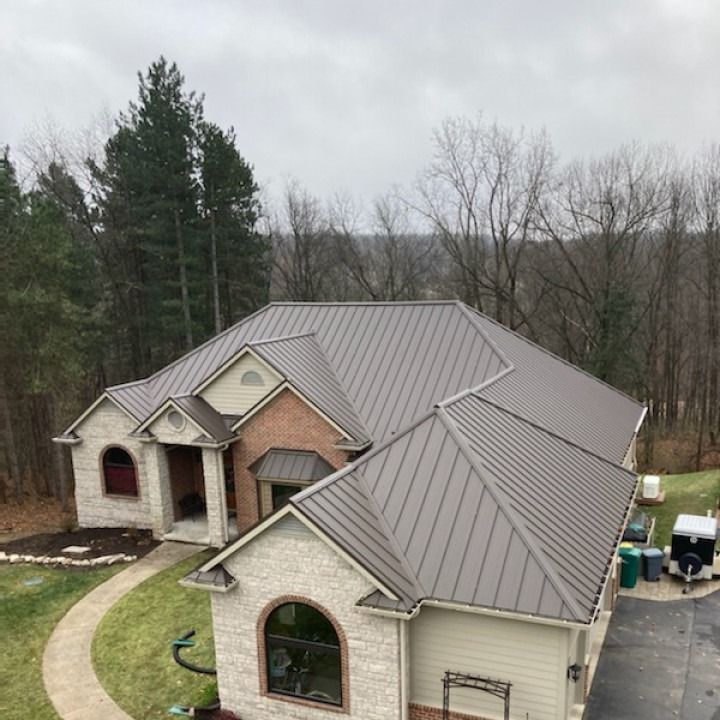 A house with a brown metal roof, brick and stone exterior, and surrounding trees under a cloudy sky.
