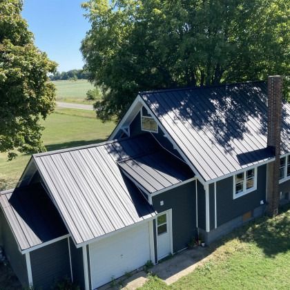 Dark house with metal roof, chimney, white garage door, and lush greenery in a sunny, rural setting.