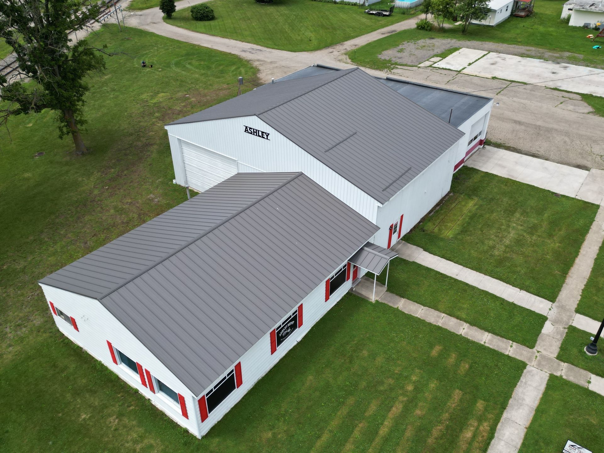 Aerial view of a white building with a grey roof and red window trim, surrounded by green grass.