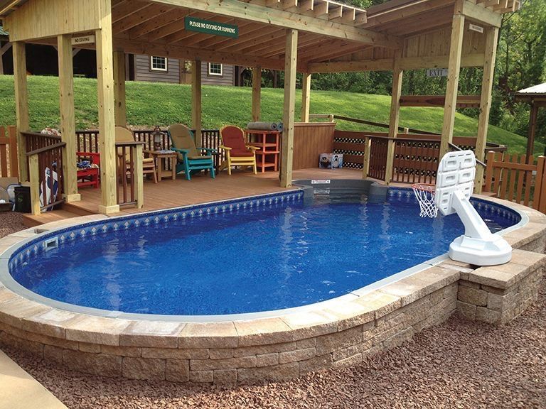 Oval-shaped pool with a basketball hoop, under a wooden pergola. Surrounded by brown landscaping and grass.