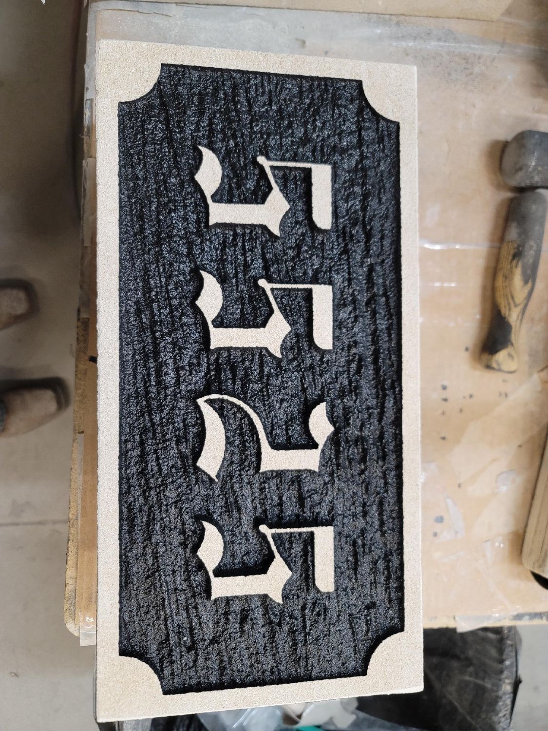 Wooden sign with black background, cream border, and three Hebrew characters.