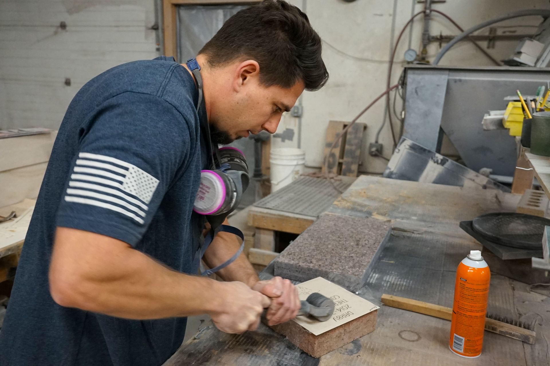 Man carving stone with a hammer and chisel in a workshop. He wears a respirator, and American flag on sleeve.