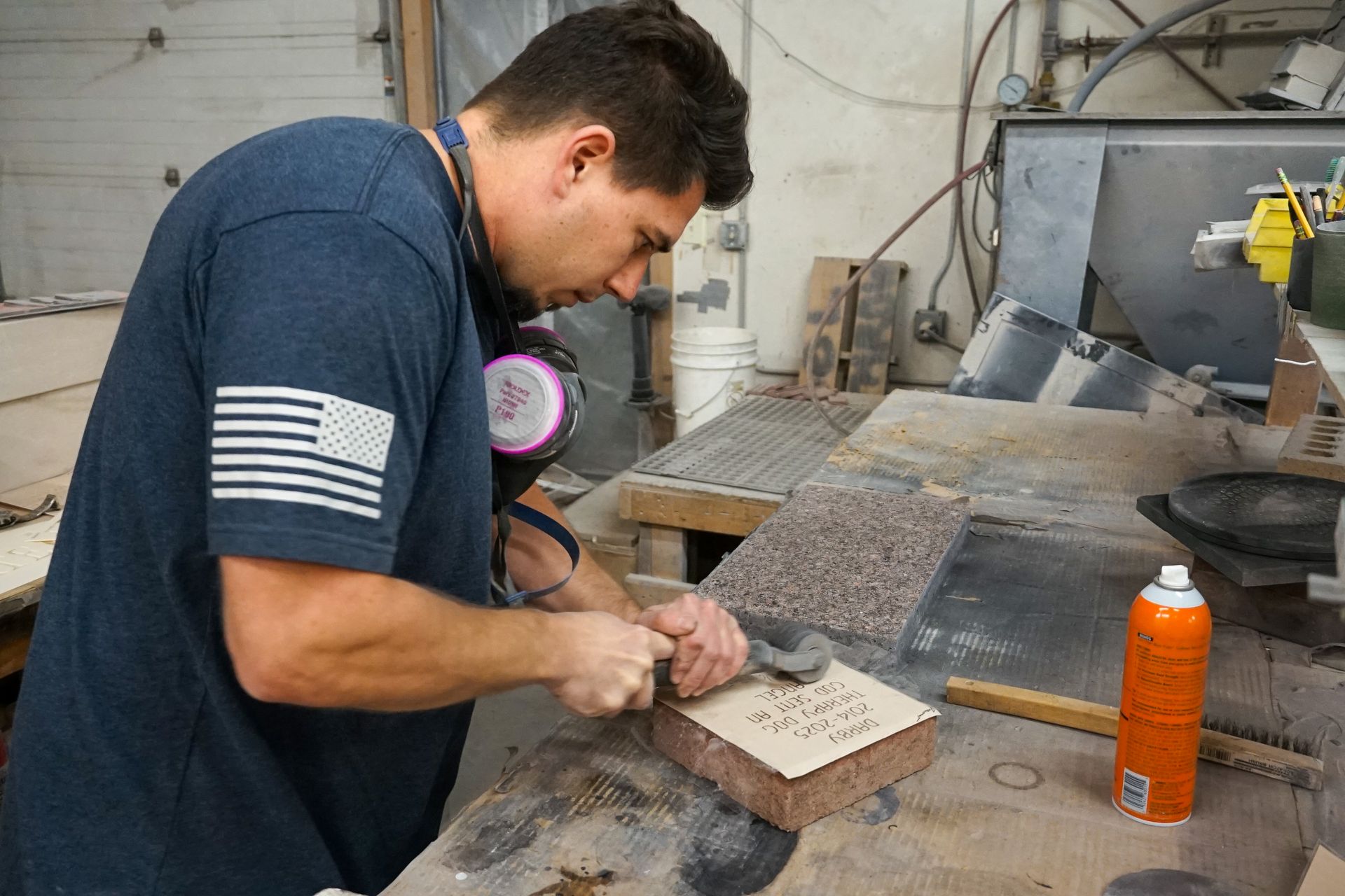 Man carving stone, wearing mask in workshop. He's using a tool on a block with holes.