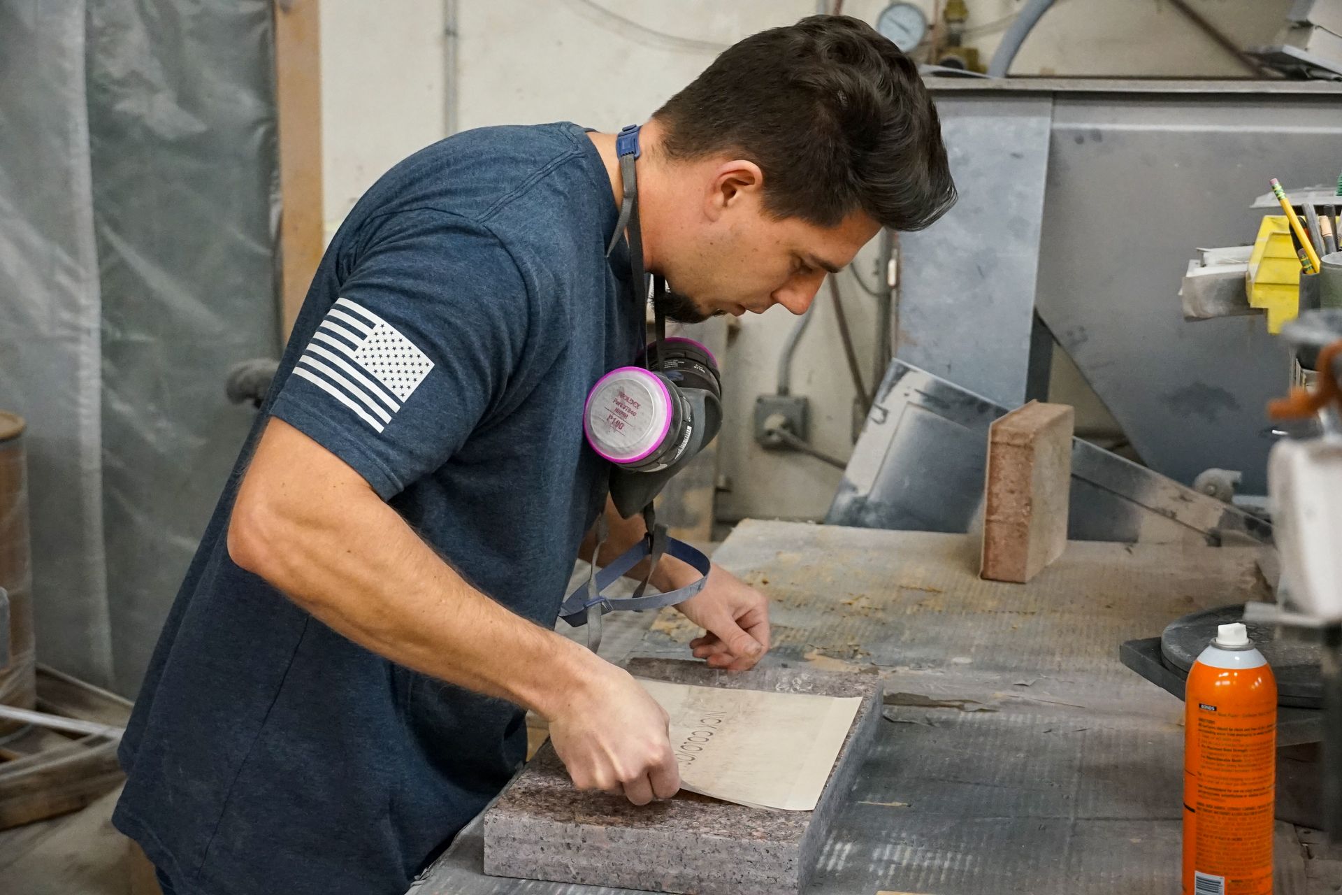 Man working at a workbench, wearing a mask and dark blue shirt with flag patch.