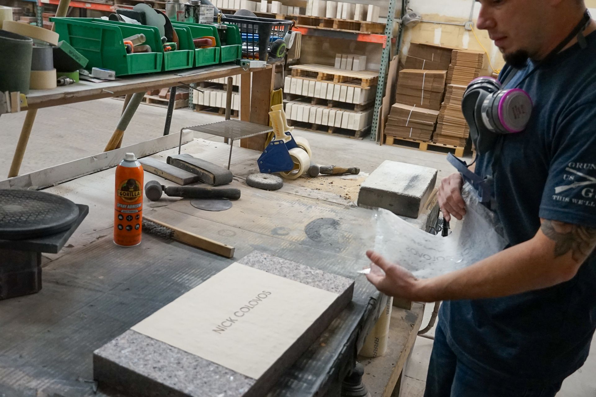 Man working with materials on a workbench, wearing a respirator. Industrial setting with supplies visible.