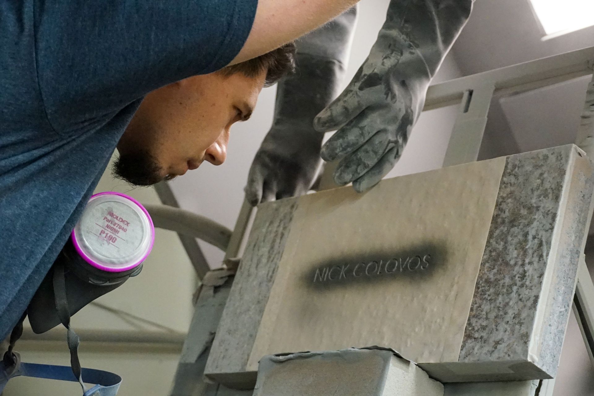 Man wearing respirator and gloves sprays lettering on stone block.