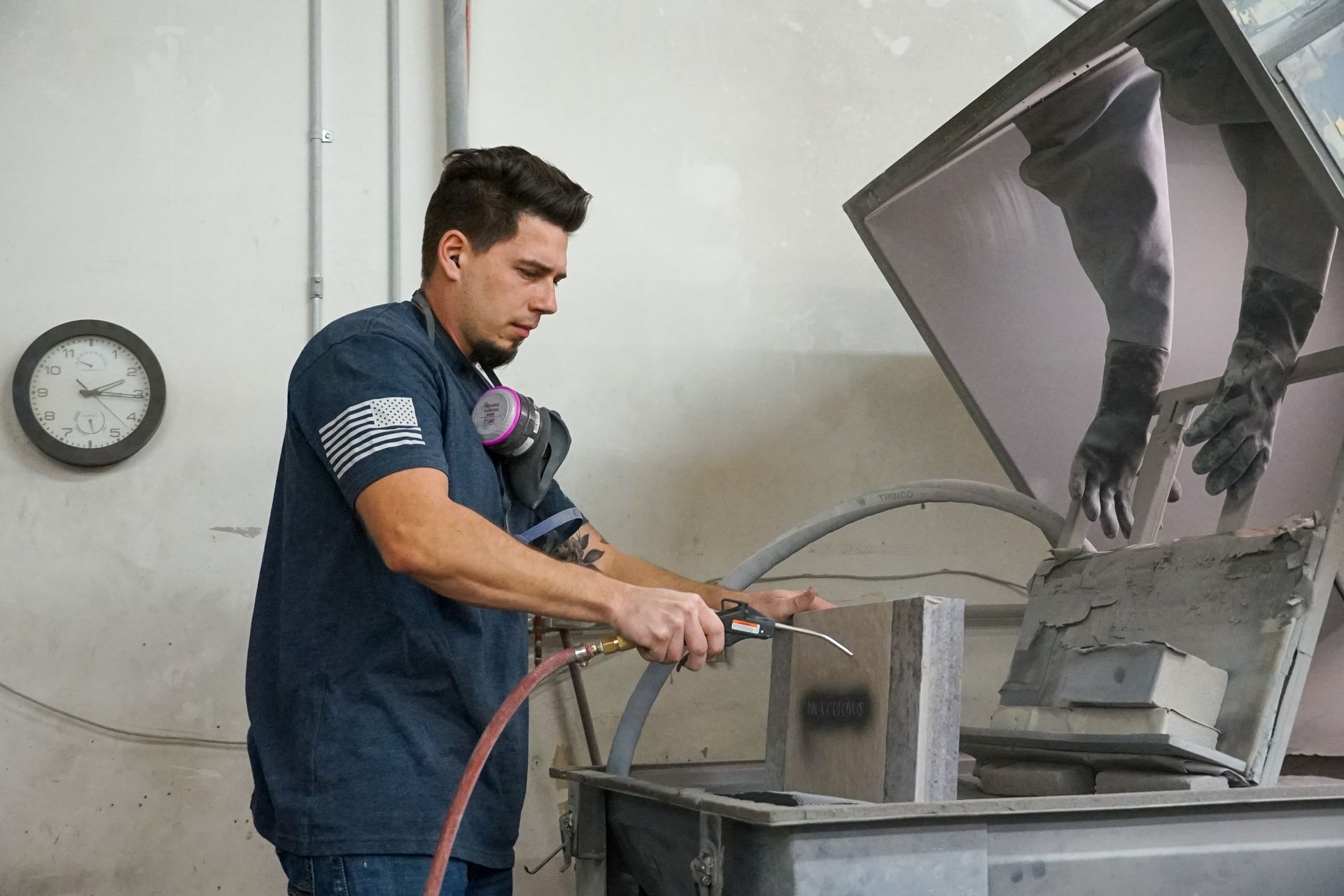 Man sandblasting a metal object inside an enclosure; he wears a respirator and gloves in a workshop.