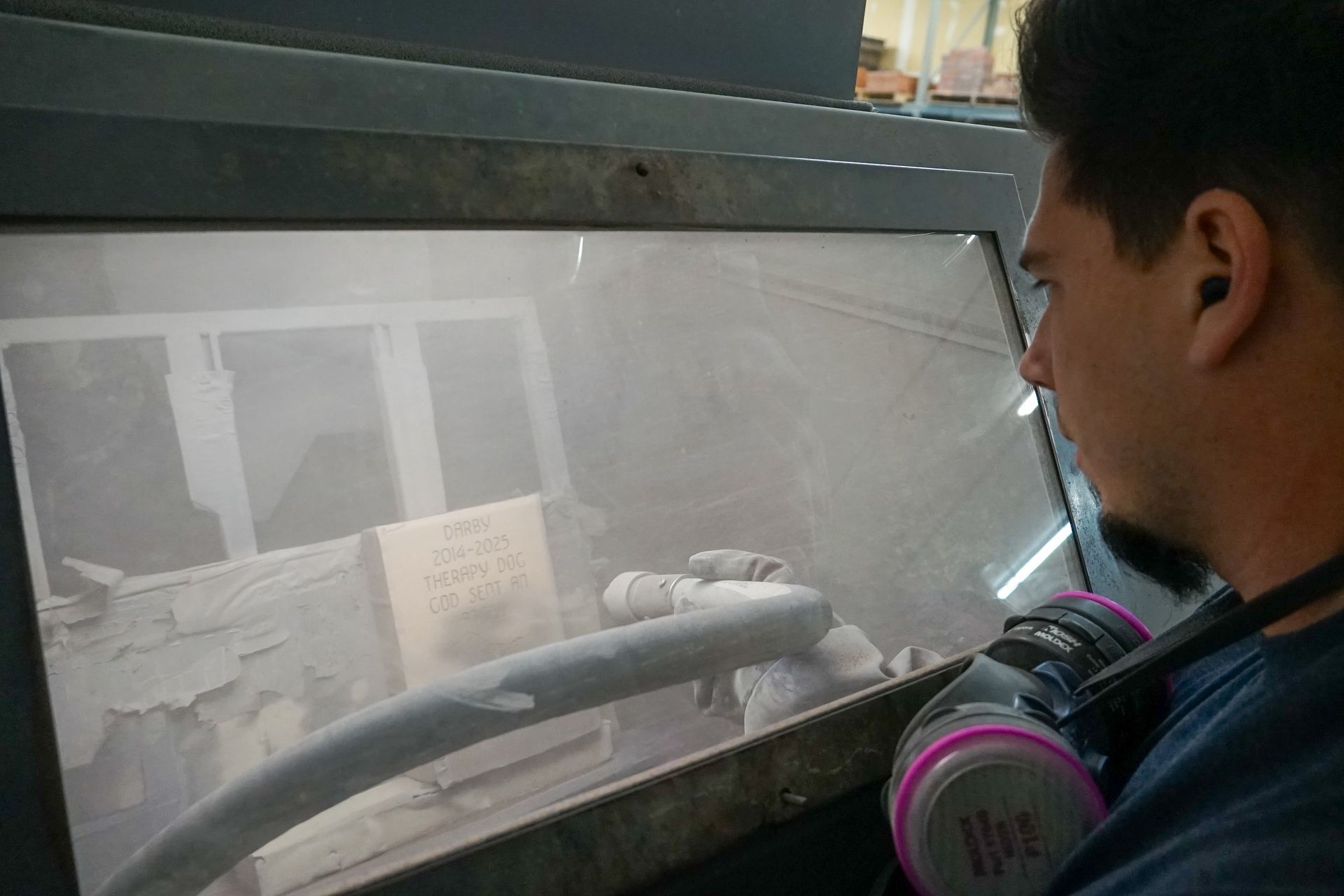 Person operating a sandblasting machine, wearing a respirator. Gray machine interior filled with dust.