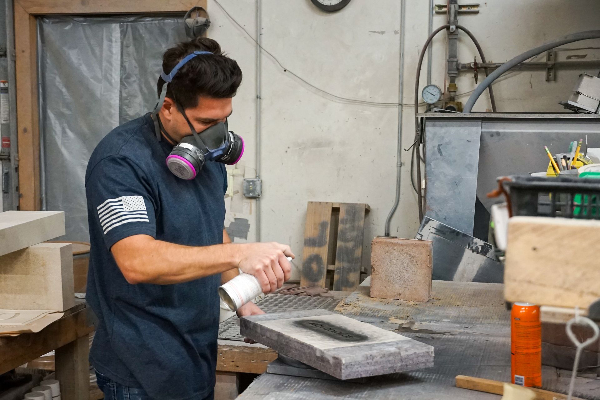 Man wearing respirator sprays a dark stone block in a workshop, grey walls and tools visible.