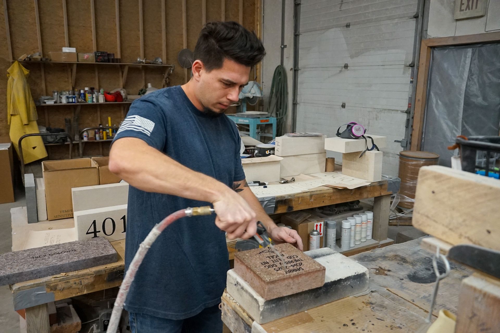 Man in blue shirt using an air hose on a brick in a workshop.