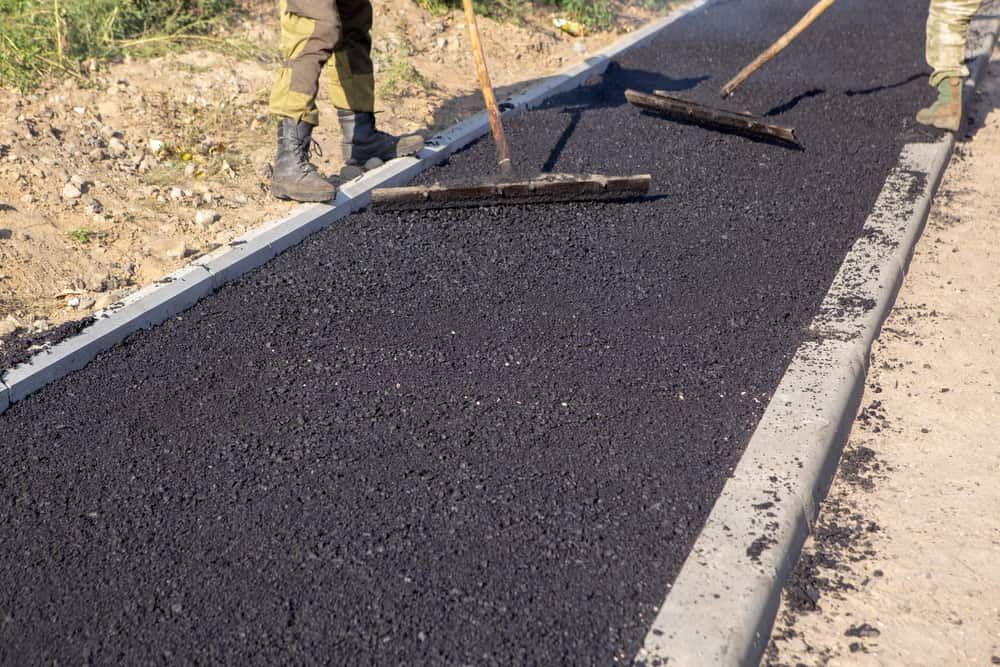 Workers use long-handled rakes to spread and smooth fresh, dark asphalt between concrete curbs — Territory Bitumen Services In Tivendale, NT