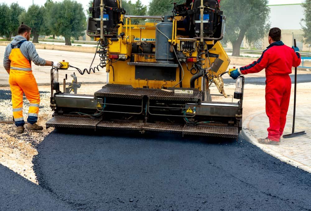 Two Men Are Standing Next To A Machine That Is Spreading Asphalt On A Road — Territory Bitumen Services In Tivendale, NT