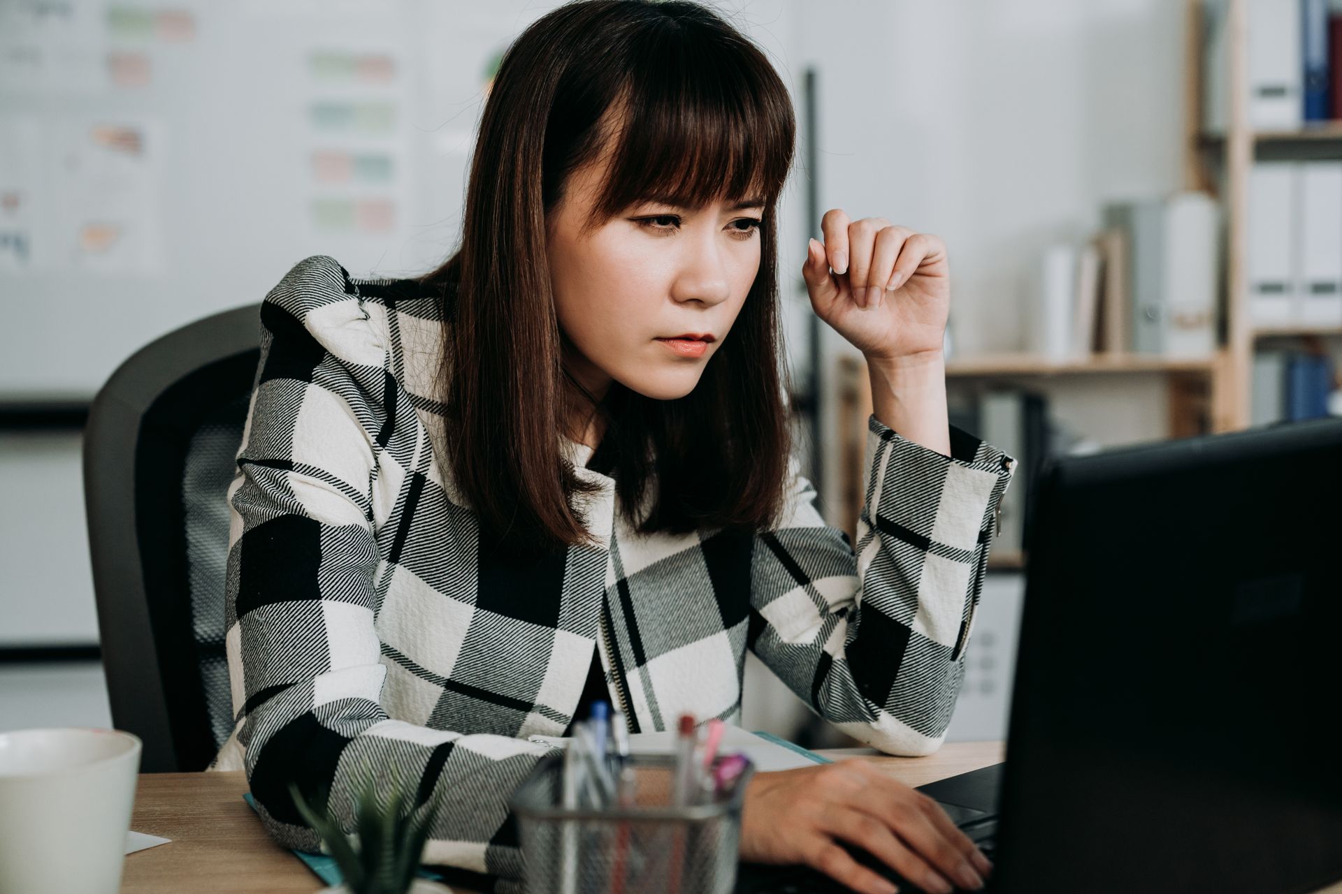 A woman is squinting while leaning forward close to a laptop monitor at an office desk.