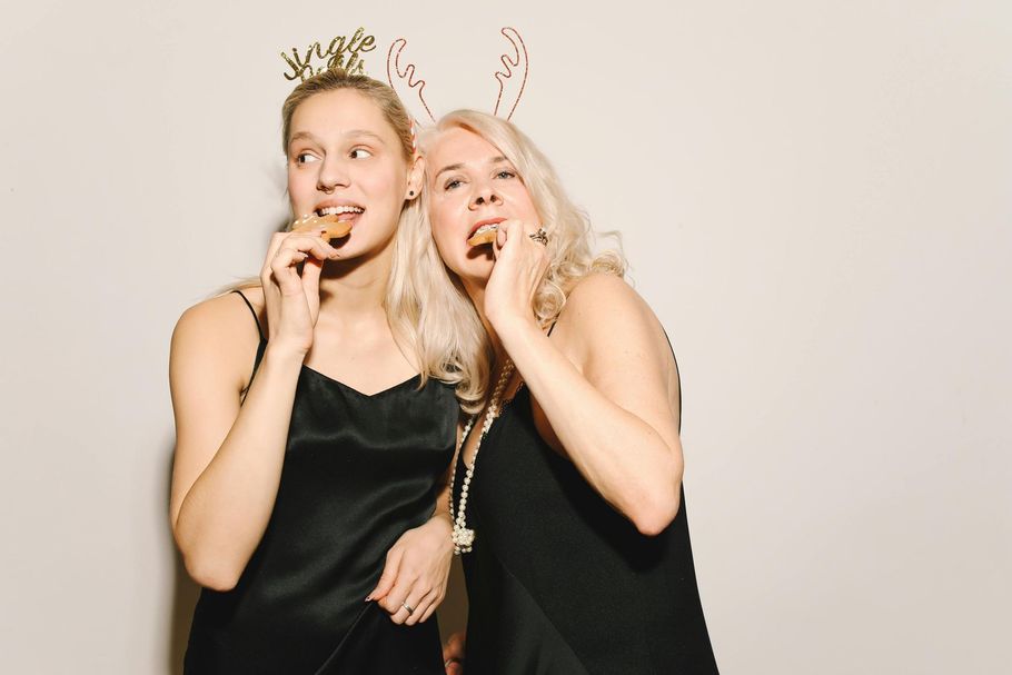 Two women in black dresses wearing party hats and eating snacks, posing together against a plain background.