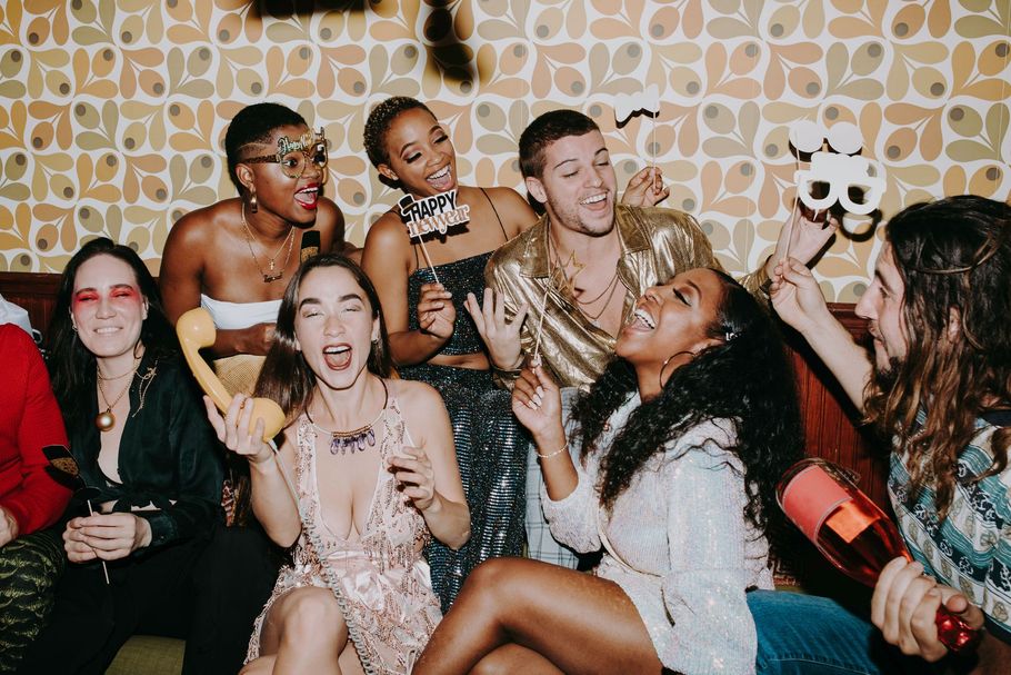 People cheering and posing at a lively party, holding drinks and smiling in front of patterned wallpaper