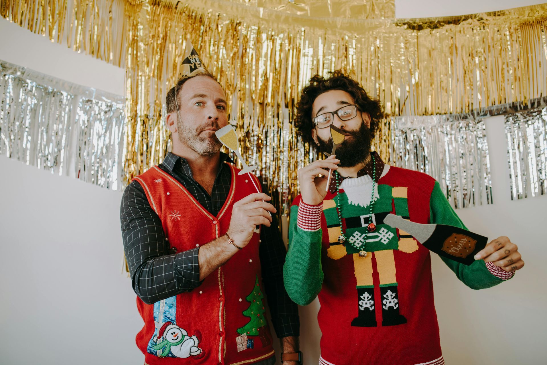 Two men in festive sweaters sing with toy microphones under gold and silver party streamers.