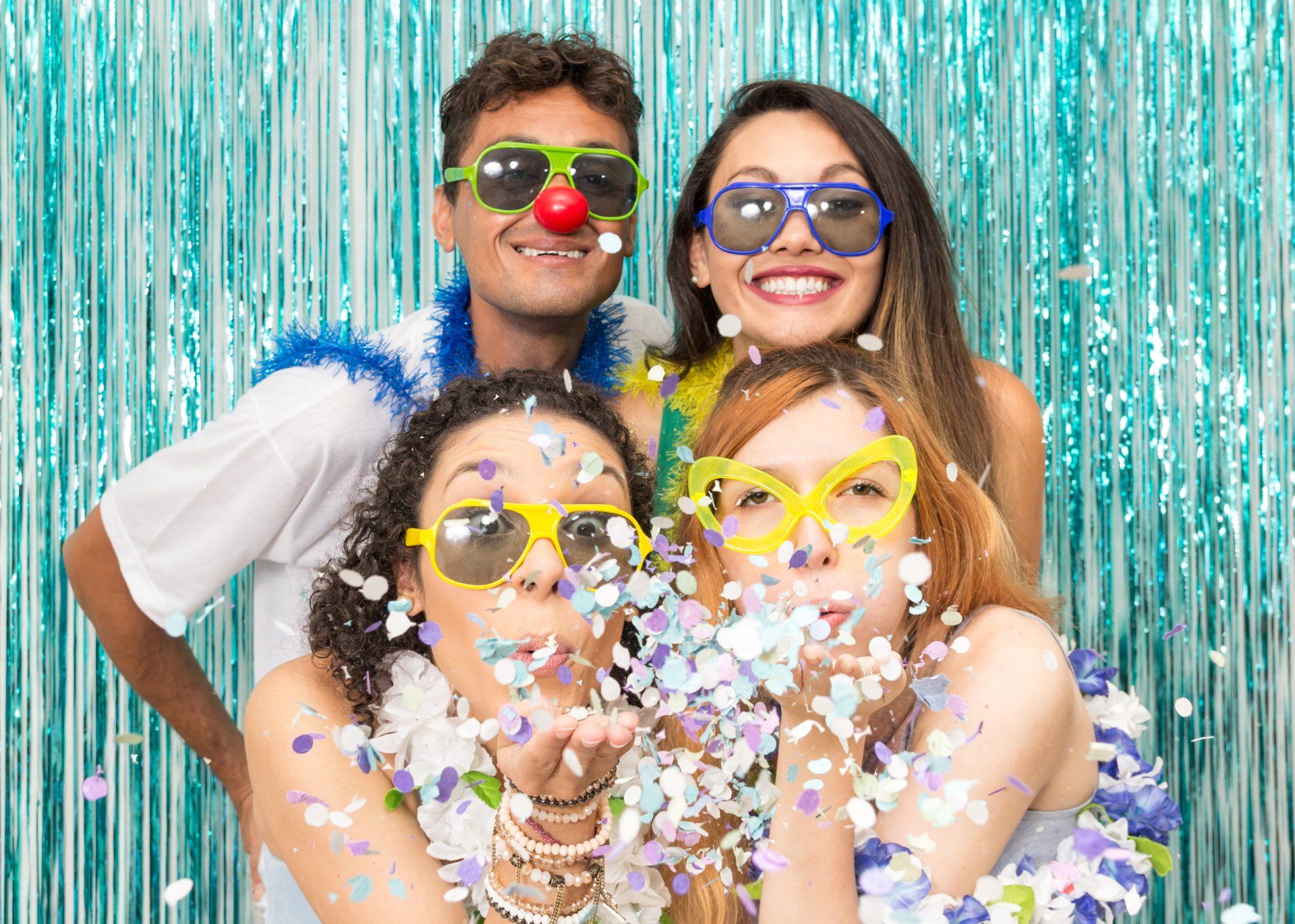 Four people in silly glasses and leis blow confetti in front of a blue tinsel backdrop.