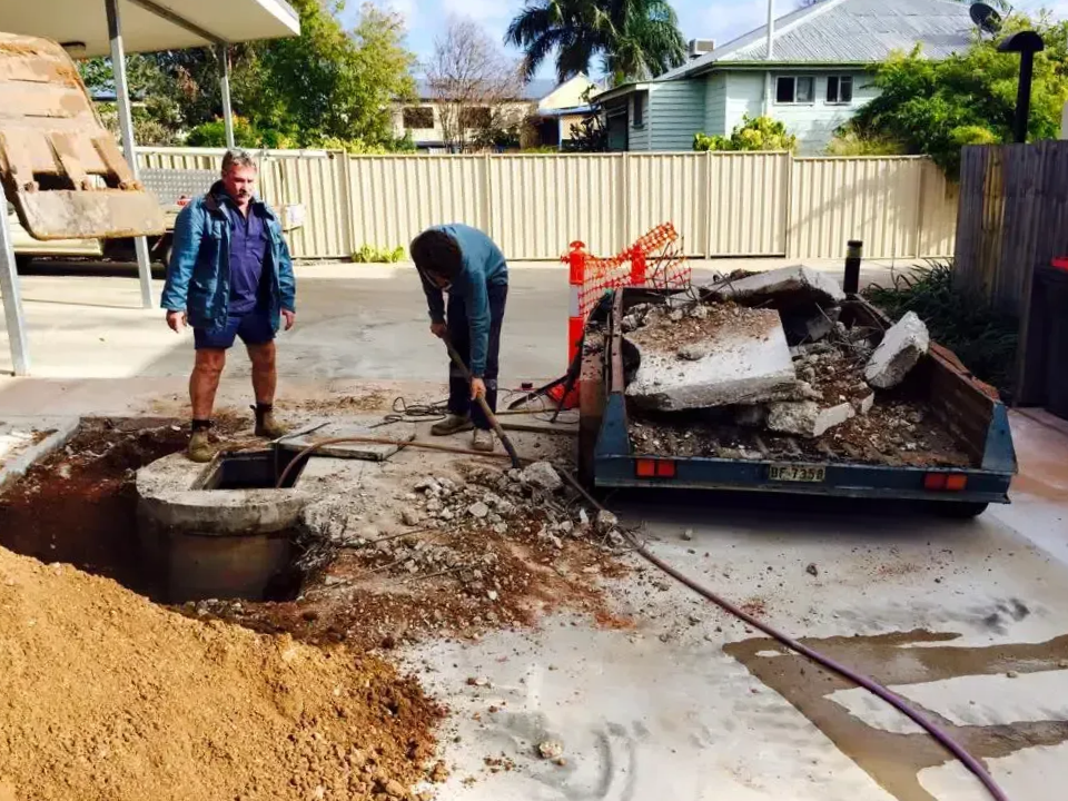 Two people work on a concrete excavation near a trailer filled with rubble in a residential driveway — Cliff Tucker Plumbing In Emerald, QLD
