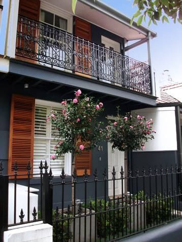 Two-story house with black facade, ornate wrought-iron balcony and fence. Tan shutters and pink flowering trees.